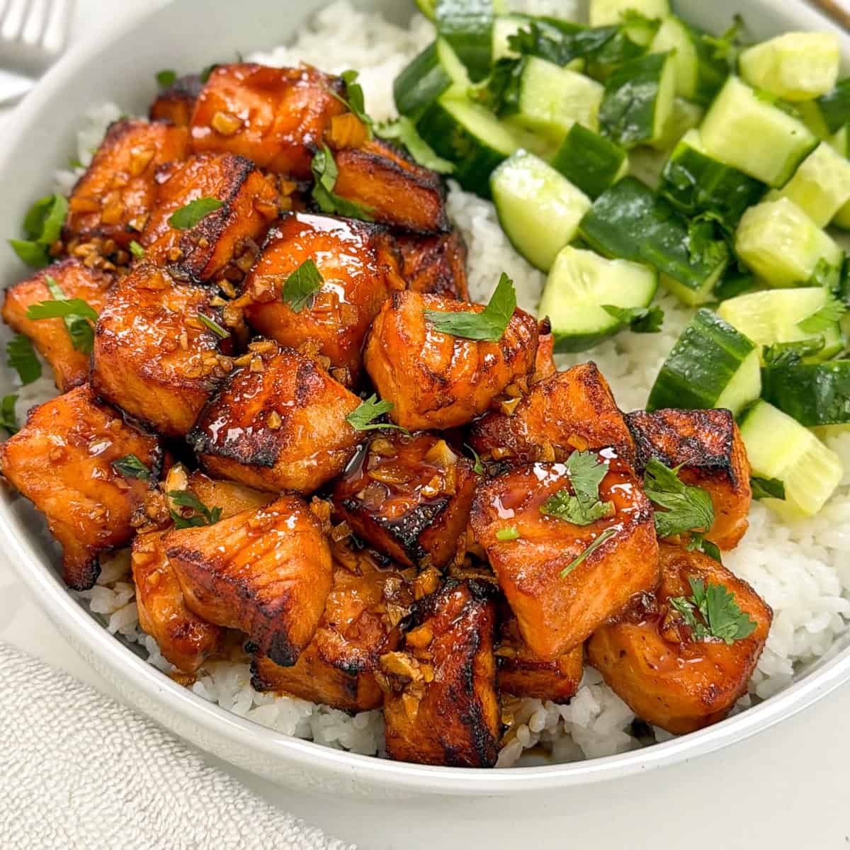A bowl of white rice topped with glazed salmon bites and diced cucumber, garnished with fresh herbs.