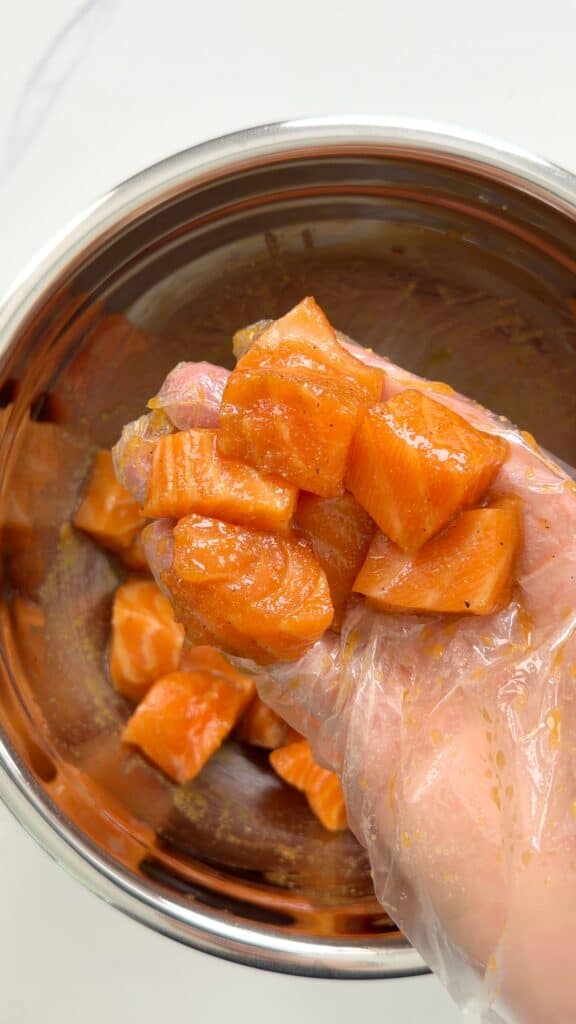 A hand wearing a clear plastic glove holds seasoned, cubed salmon over a metal mixing bowl.