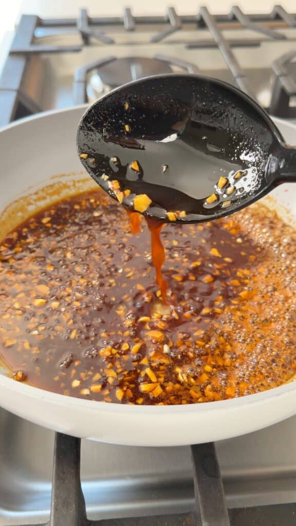 A black spoon drizzling dark brown, bubbling sauce with minced garlic into a white pan on a stovetop.