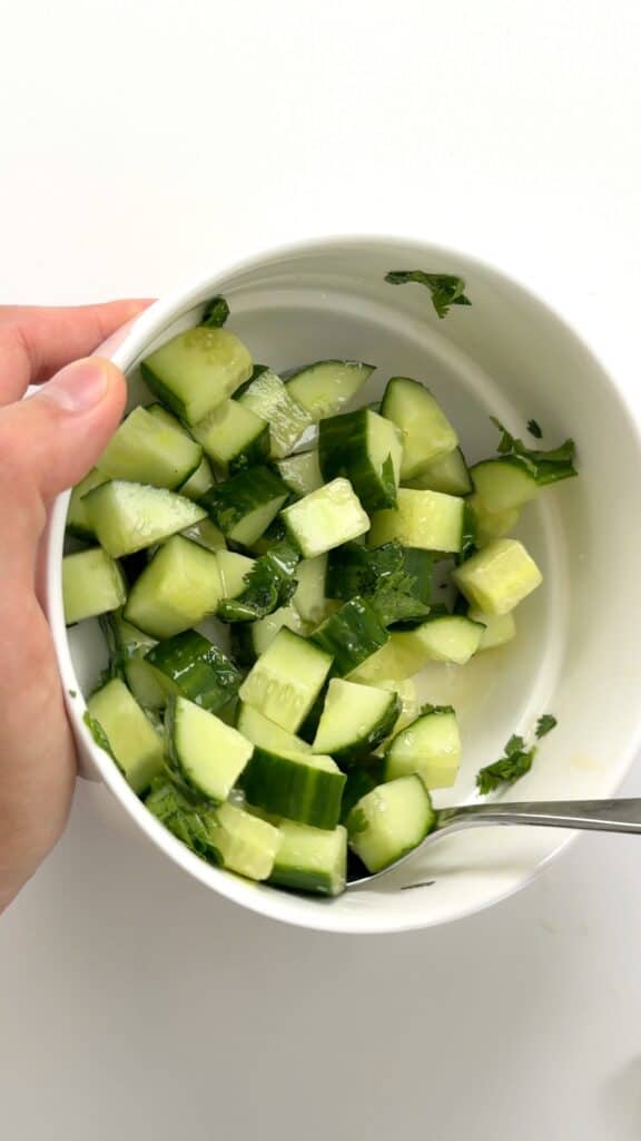 A hand holds a white bowl filled with chopped cucumbers and herbs, with a spoon resting inside.