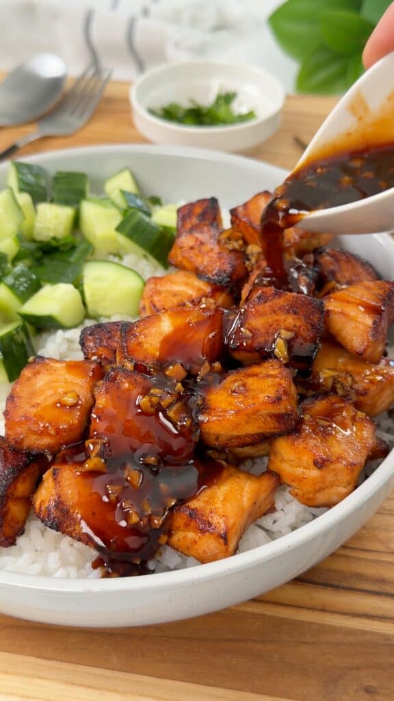 A bowl of white rice topped with glazed salmon bites and diced cucumber, with a spoonful of sauce being poured over the fish.