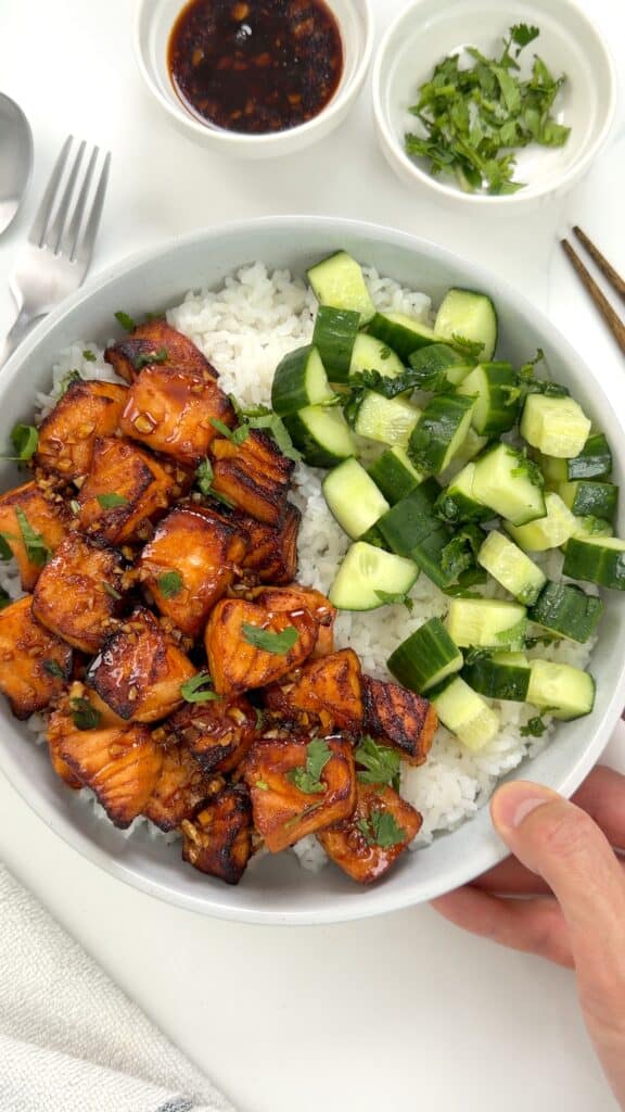 A bowl of white rice topped with glazed salmon cubes and chopped cucumbers, served with side dishes of dipping sauce and cilantro.
