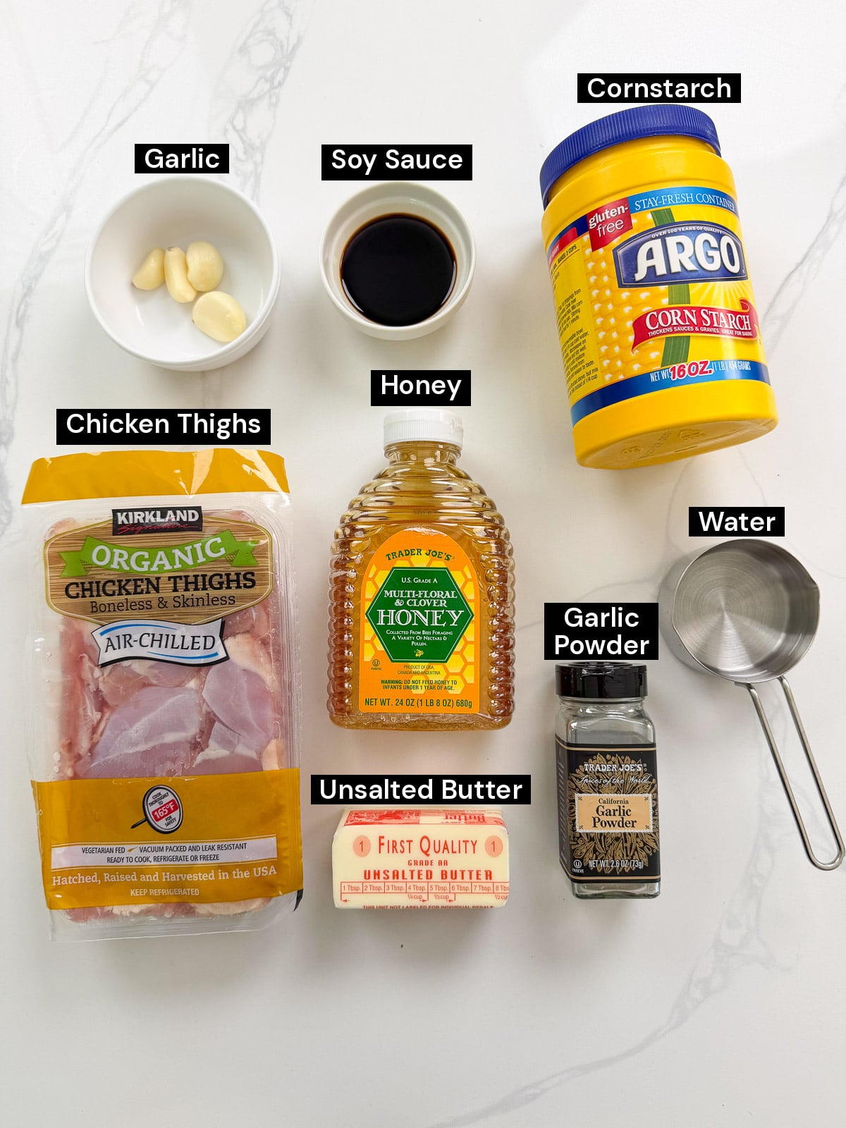 Ingredients for a recipe arranged on a white surface with labels: garlic, soy sauce, cornstarch, chicken thighs, honey, water, garlic powder, and unsalted butter.