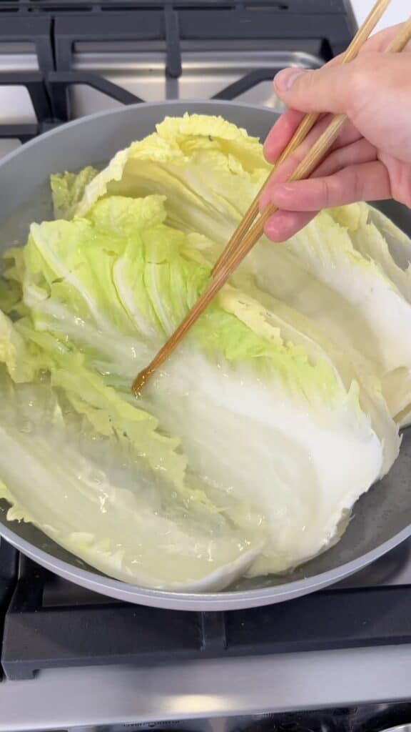 A hand using wooden chopsticks to submerge napa cabbage leaves in a pan of simmering water.