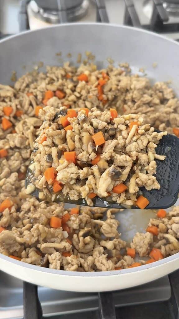 A spatula lifting a mixture of ground meat, diced carrots, and chopped mushrooms from a pan.