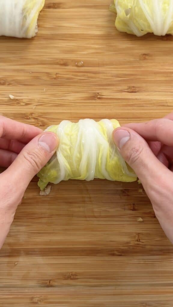 Hands rolling a cabbage leaf around a filling on a wooden cutting board.