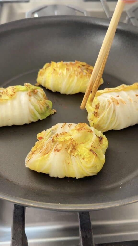 Wooden chopsticks flip a seared cabbage roll in a frying pan containing three other cabbage rolls.