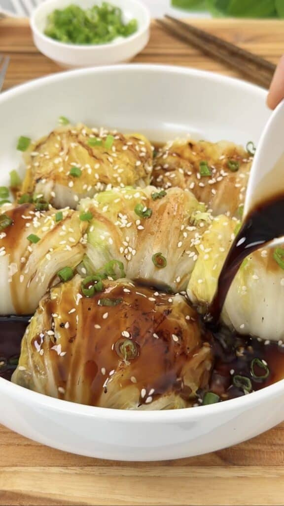 A white bowl of cabbage rolls topped with sesame seeds and green onions, with dark sauce being poured over them from a spoon.
