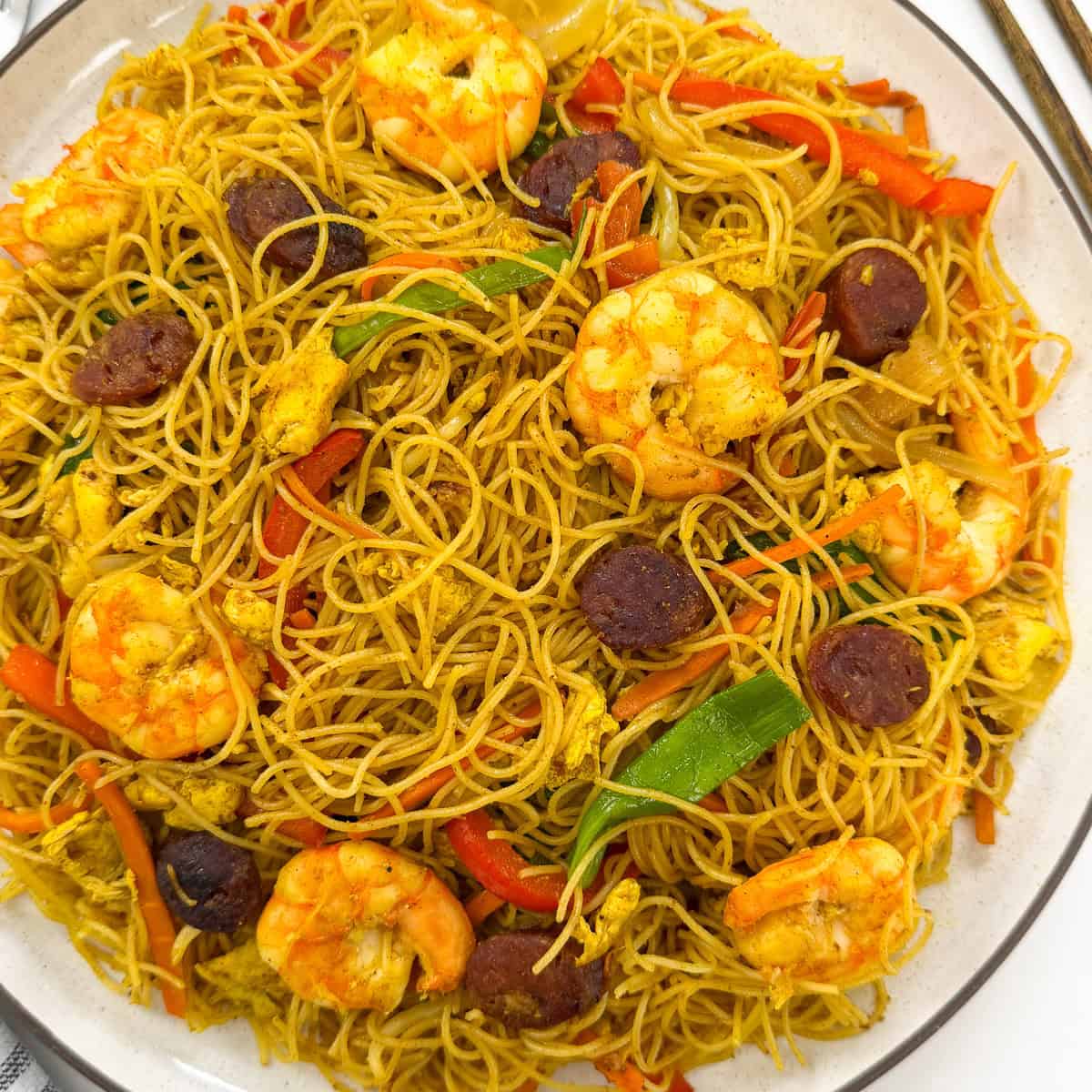 Singapore-style rice vermicelli stir-fried with shrimp, sliced sausage, egg, and peppers on a white plate.