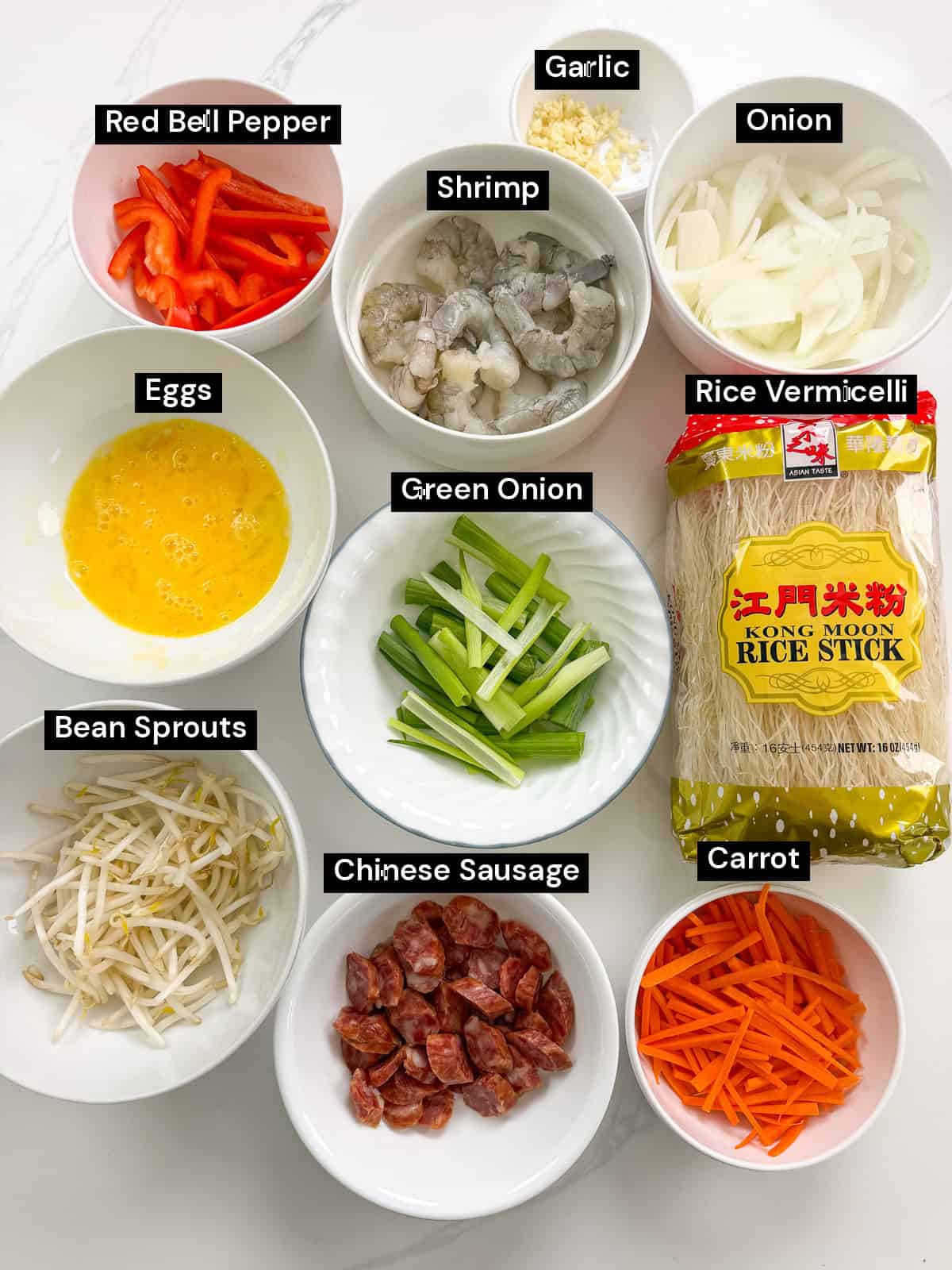 Ingredients for a stir-fry laid out in bowls, including shrimp, eggs, bean sprouts, sliced red pepper, onion, green onion, Chinese sausage, carrot, garlic, and a package of Kong Moon rice sticks.