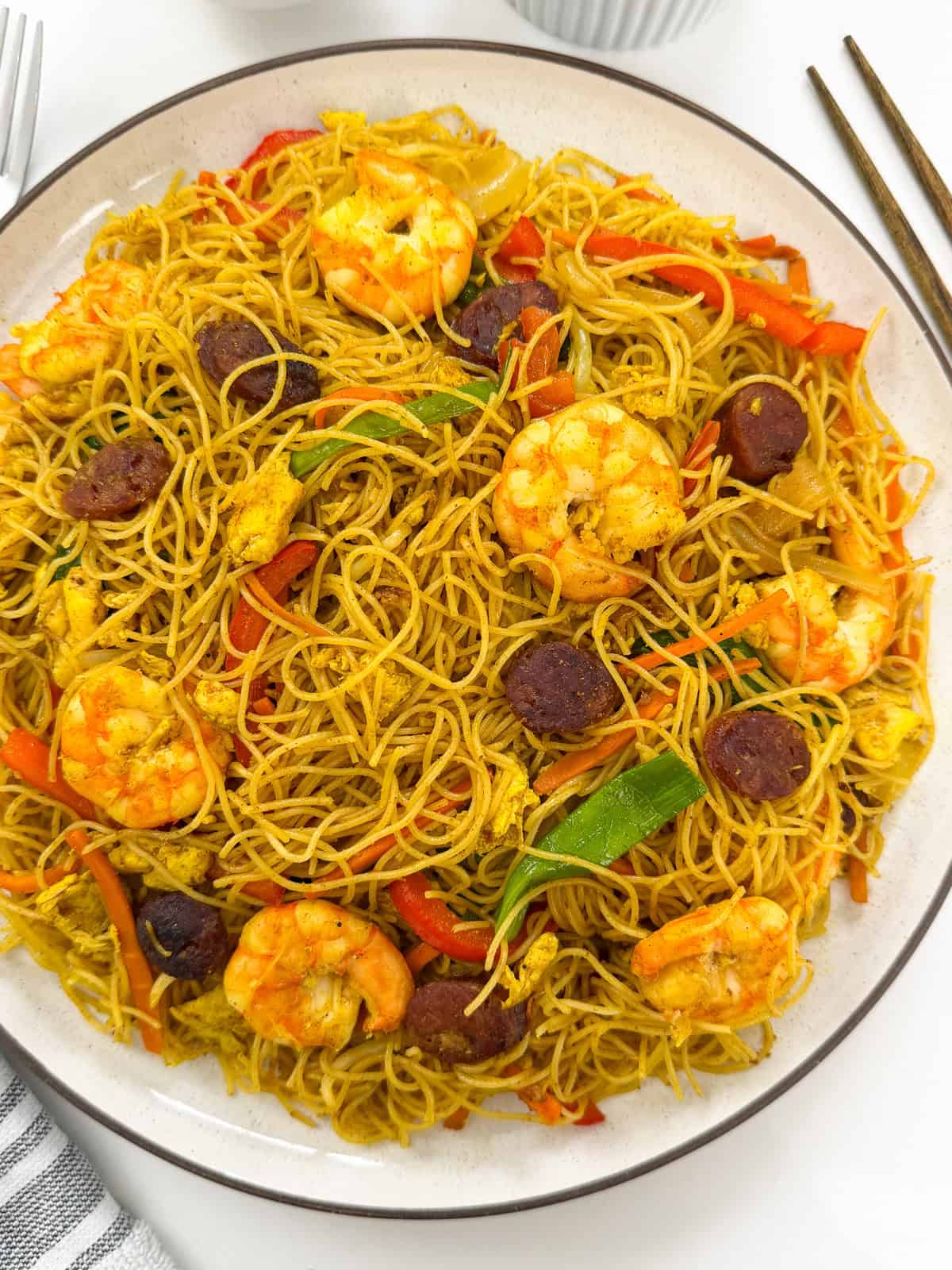 A plate of Singapore-style rice noodles stir-fried with shrimp, sliced sausage, egg, and vegetables, served with a pair of chopsticks.