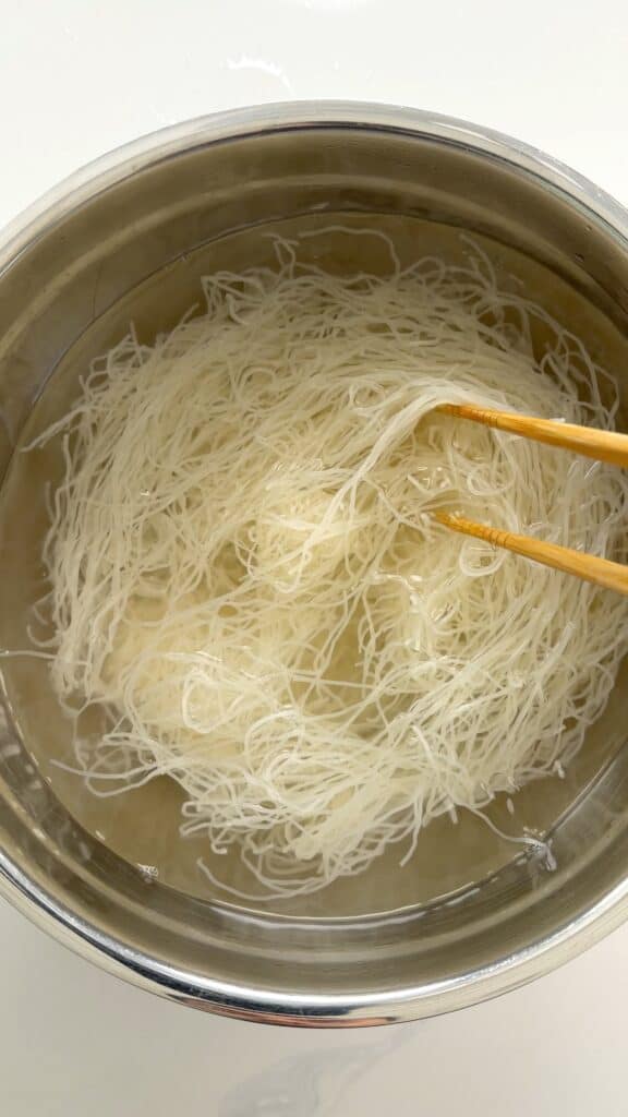 Thin rice noodles soaking in a metal pot of water, lifted by wooden chopsticks.