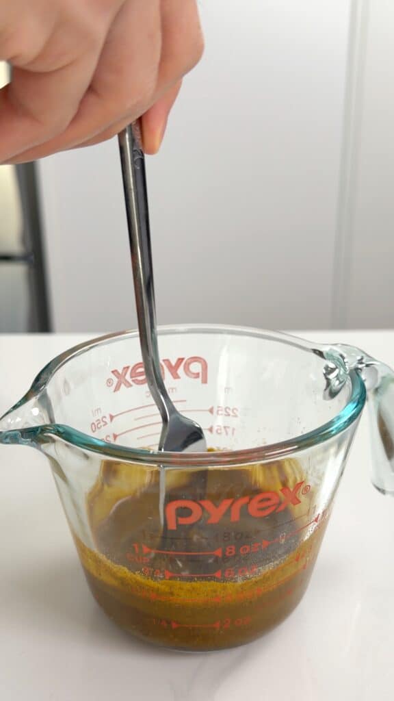 A hand uses a spoon to stir a dark brown liquid mixture inside a glass measuring cup.