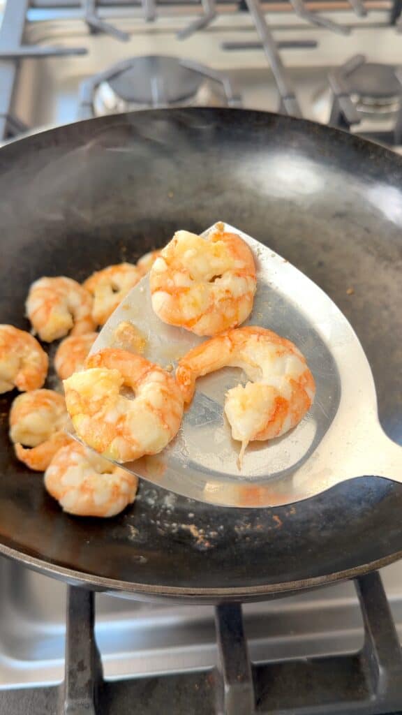 Two cooked shrimp on a metal spatula held over a frying pan containing more shrimp.