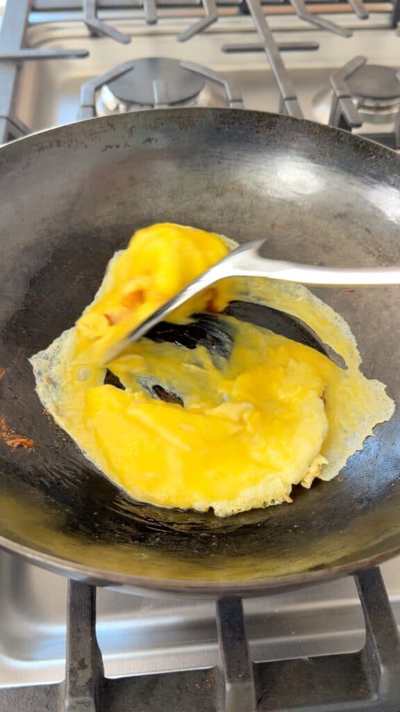 A metal spatula flips beaten eggs cooking in a dark frying pan on a stovetop.