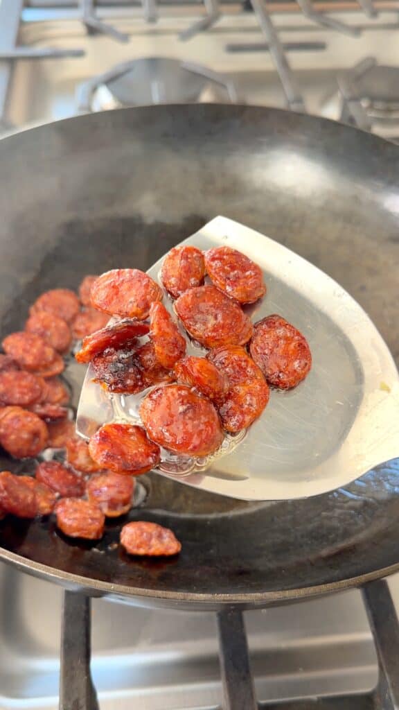 A metal spatula holds a portion of fried chorizo slices over a skillet.