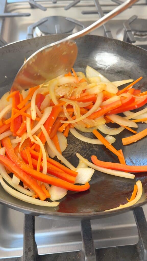 A metal spatula stirs sliced red peppers, carrots, and onions in a black skillet on a stovetop.