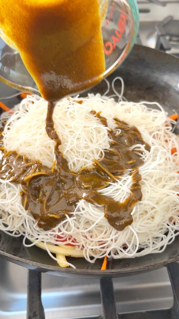 Dark sauce being poured from a measuring cup onto a wok filled with thin rice noodles and shredded carrots.