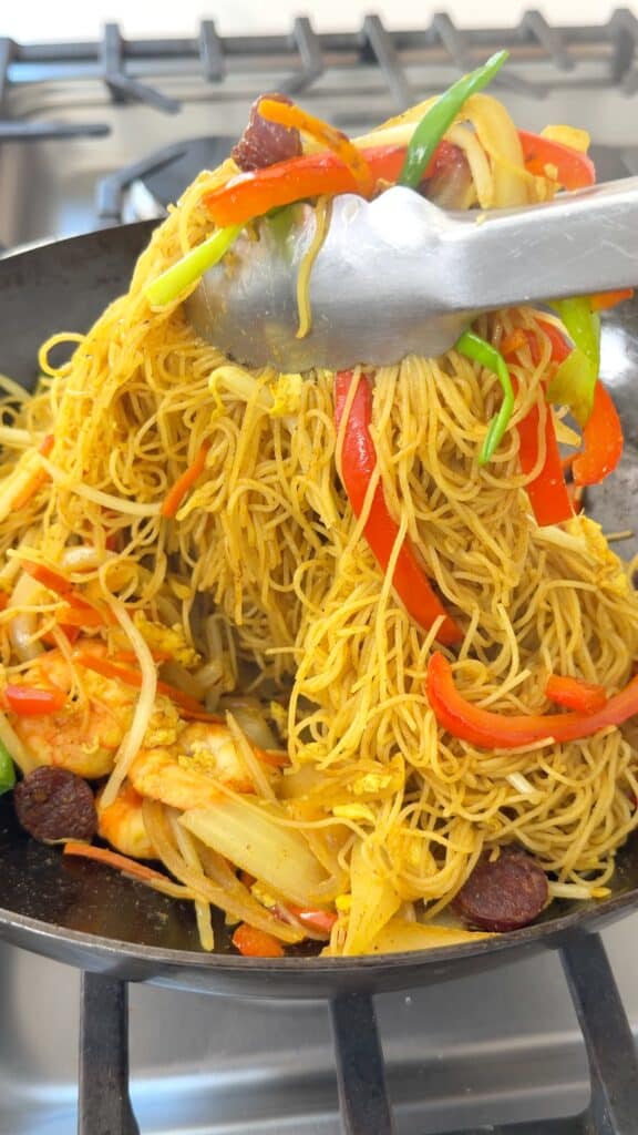 Metal tongs lift stir-fried rice vermicelli noodles mixed with shrimp, sliced red peppers, green onions, and sausage in a wok.