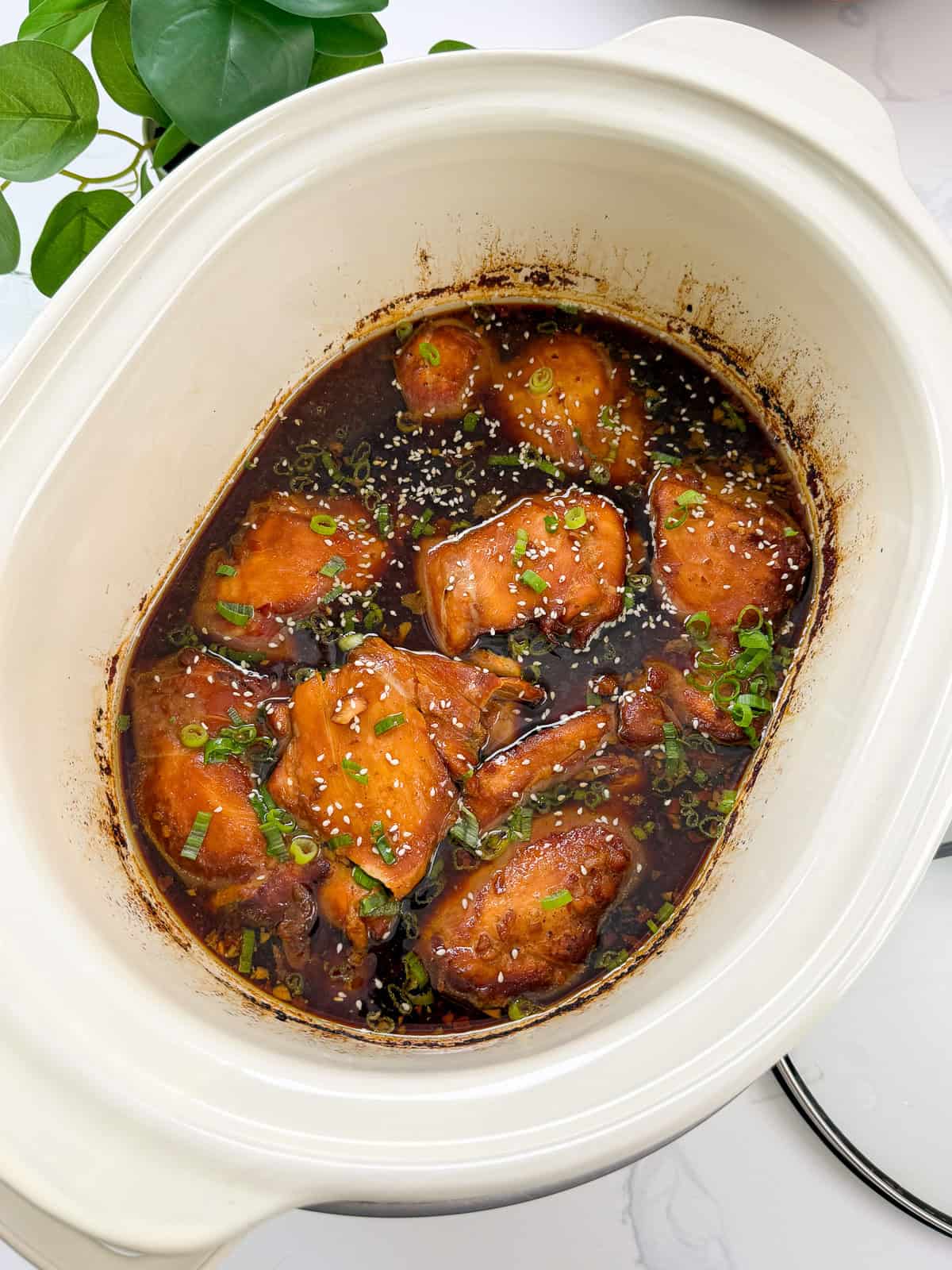 Glazed chicken thighs in a dark savory sauce, garnished with sesame seeds and sliced green onions in a slow cooker.