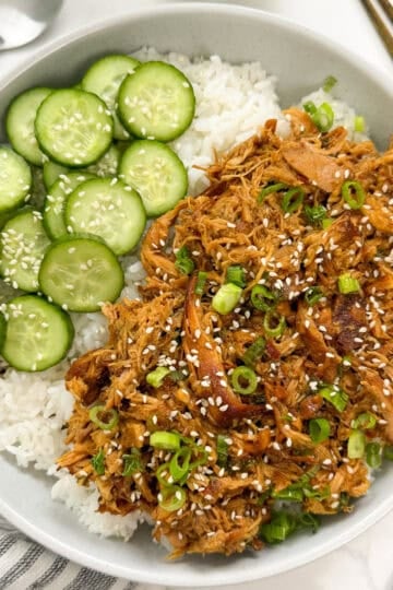 A bowl of white rice topped with shredded chicken, sliced cucumbers, sesame seeds, and green onions.