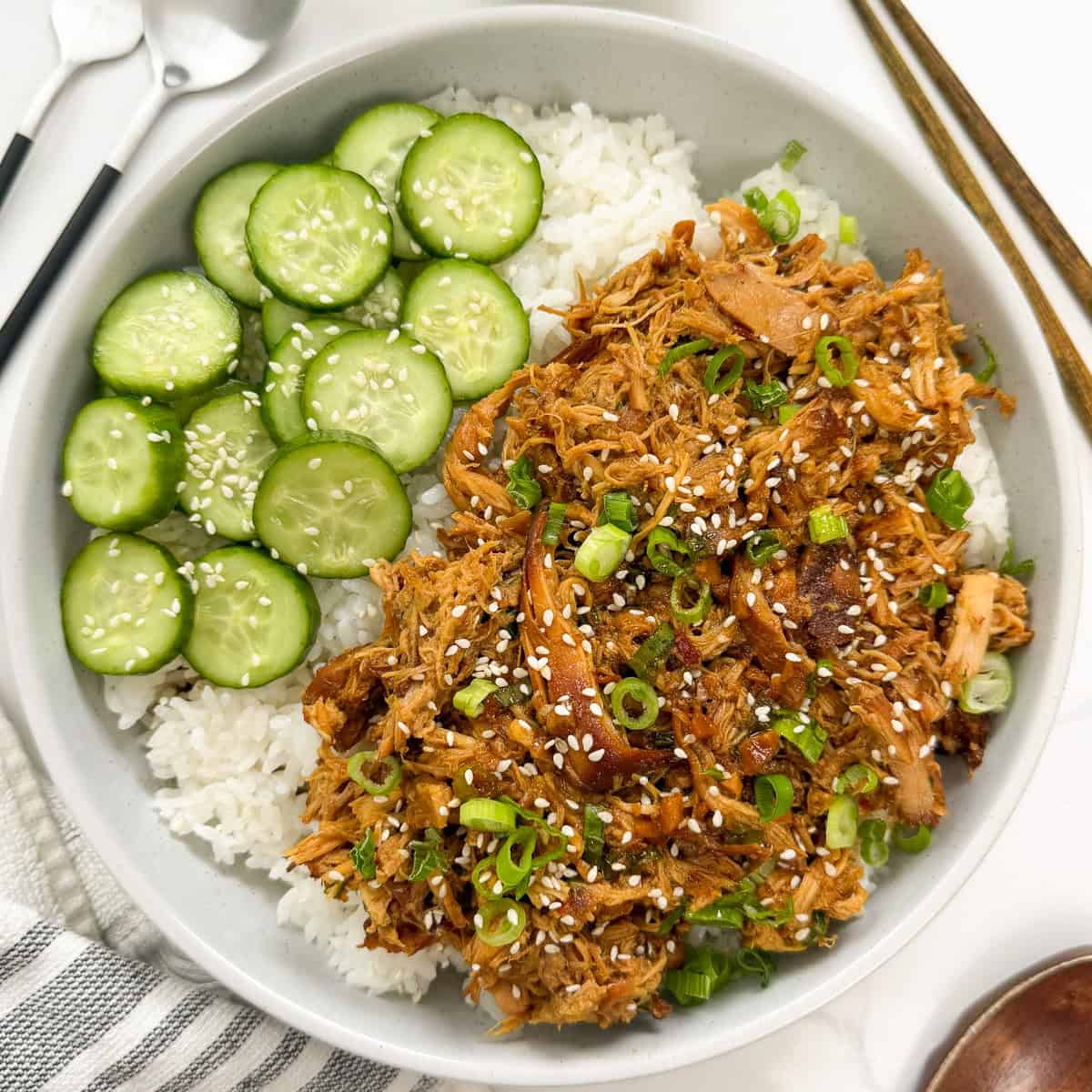 A bowl of white rice topped with shredded chicken, sliced cucumbers, sesame seeds, and green onions.