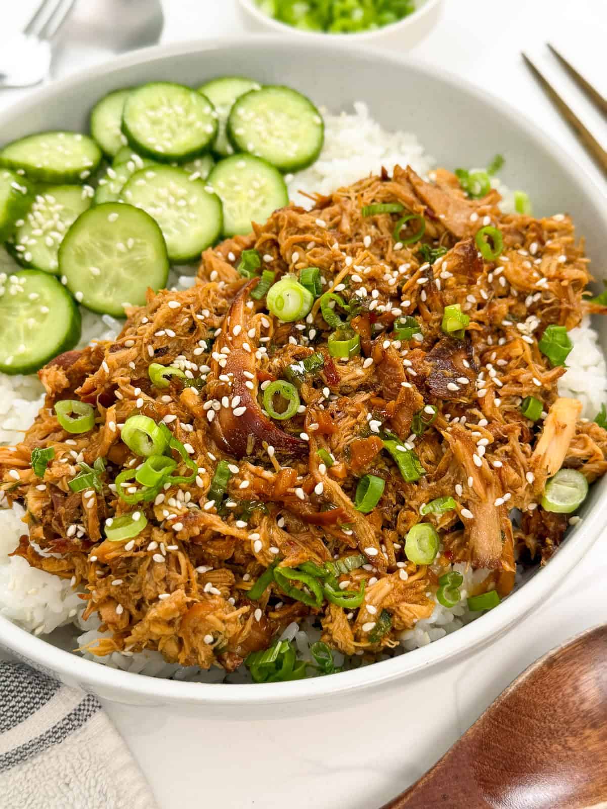 A bowl of shredded meat over white rice, topped with sesame seeds and sliced green onions, served with a side of cucumber slices.
