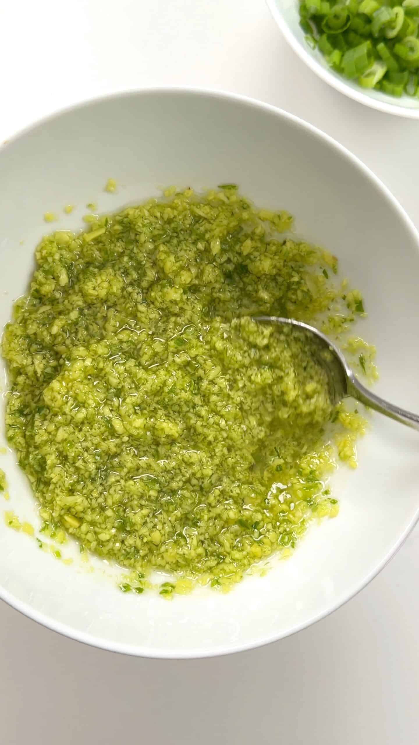 A white bowl containing a green herb and garlic oil mixture being stirred with a spoon, with a small bowl of chopped green onions nearby.