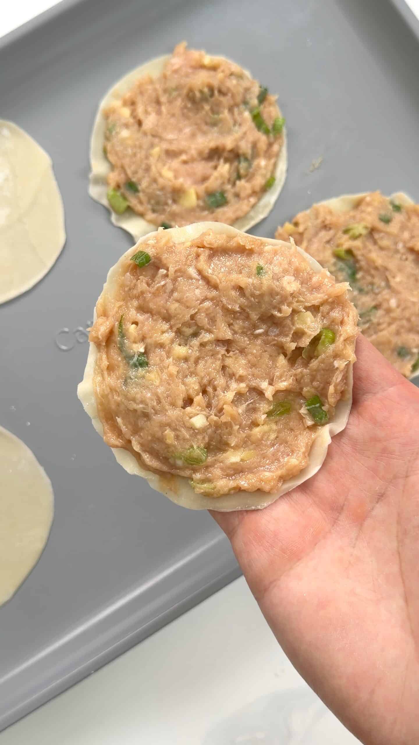 A hand holds a dumpling wrapper topped with raw meat filling and green onions, over a baking sheet with more filled wrappers.