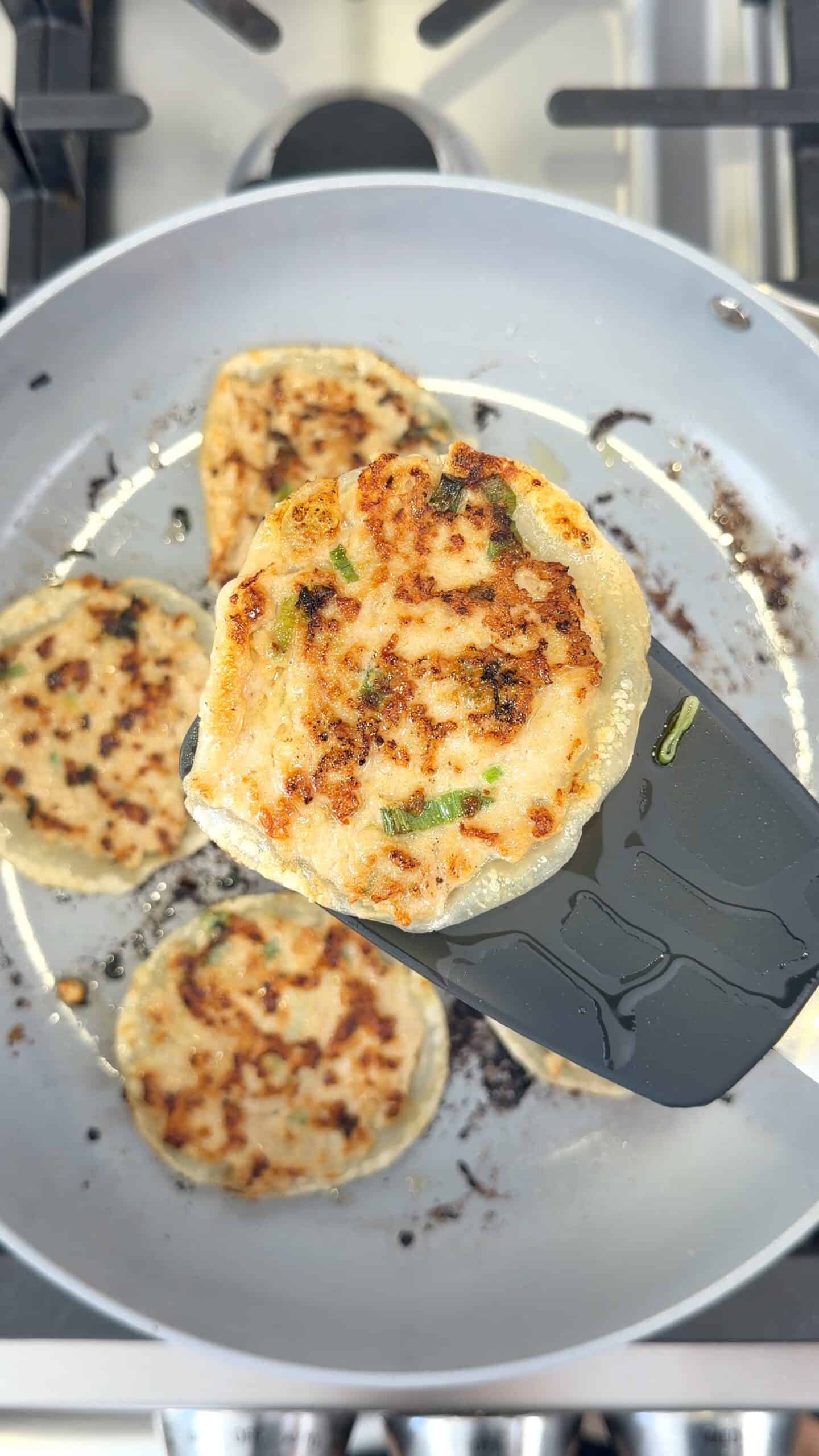 A spatula lifts a browned savory pancake with visible green onions from a frying pan containing three other similar pancakes.
