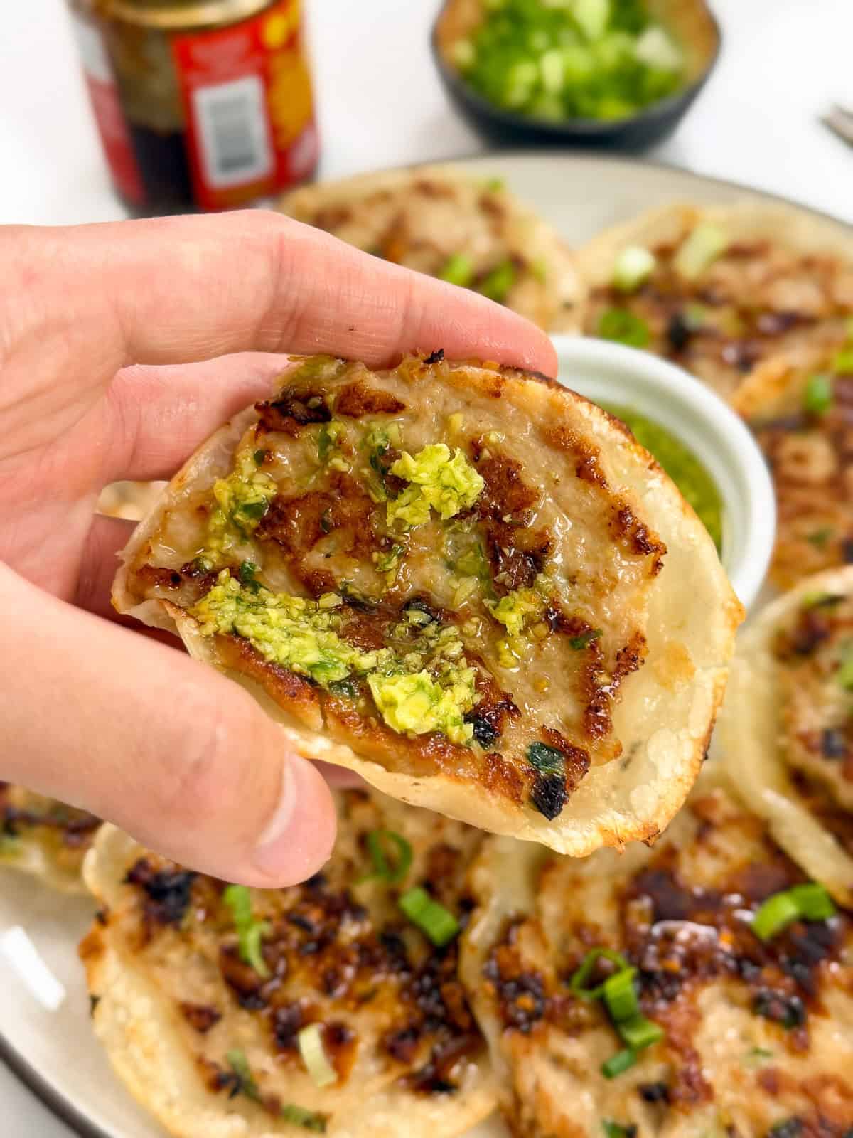 A hand holds a half-eaten, crispy dumpling filled with meat and topped with green scallion sauce. More dumplings and a small bowl of dipping sauce are on a plate in the background.