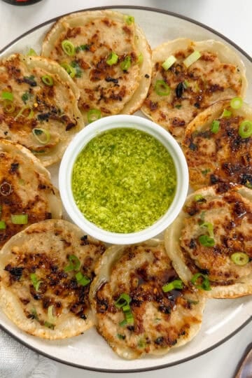 A plate of crispy pan-fried dumplings arranged around a small bowl of green dipping sauce, garnished with sliced scallions. Chopsticks and a napkin are beside the plate.