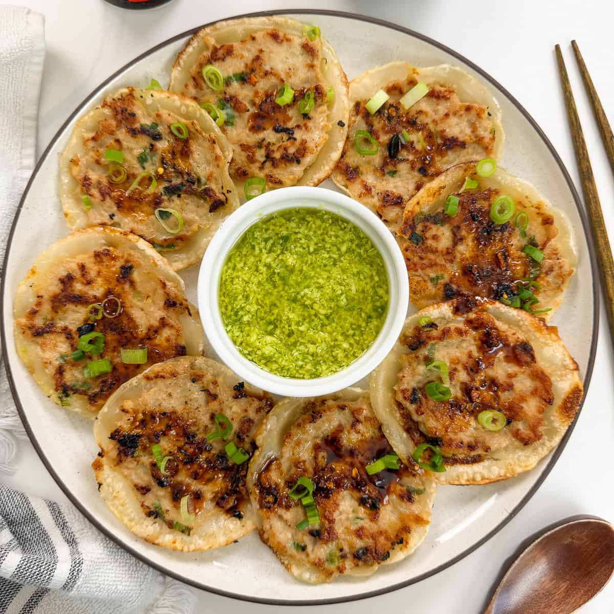 A plate of crispy pan-fried dumplings arranged around a small bowl of green dipping sauce, garnished with sliced scallions. Chopsticks and a napkin are beside the plate.