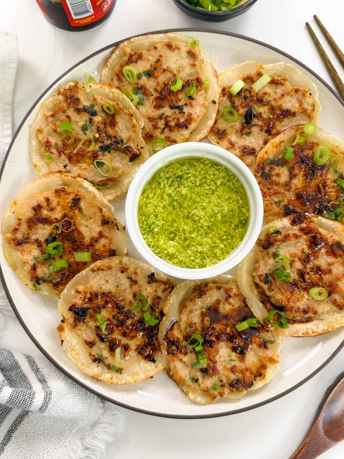 A plate of pan-fried dumplings garnished with chopped green onions surrounds a small bowl of green dipping sauce. Chopsticks and a striped napkin are nearby on a white surface.